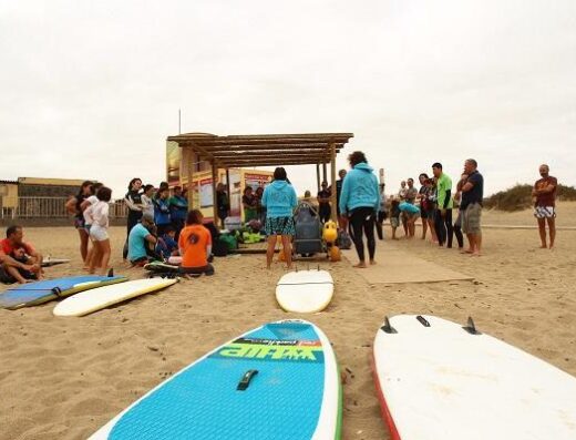 Grupo de alumnos con los instructores. Pasarela de madera y silla anfibia como elementos que permiten la accesibilidad a la actividad acuática. Imagen cedida por Escuela Fuerte Tribu Surf, Fuerteventura.