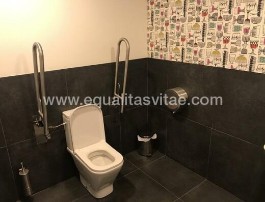 Baño adaptado con barras de apoyo y espacio de transferencia.