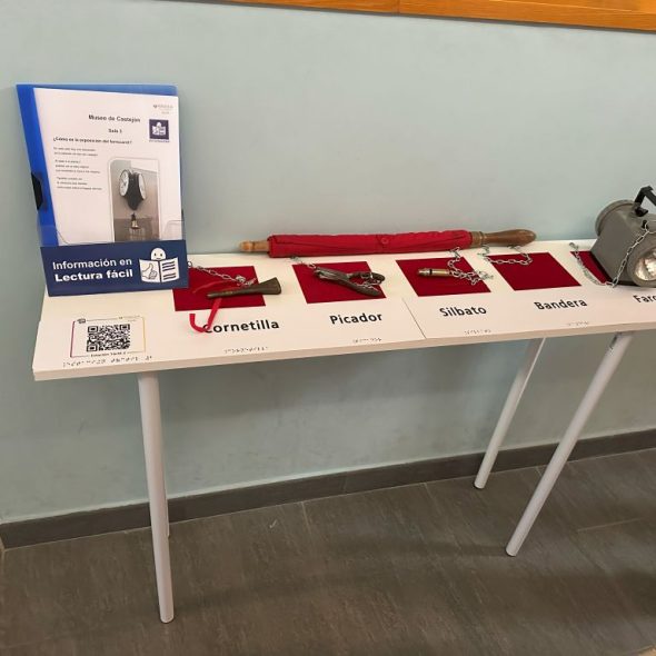 Imagen de una estación táctil con materiales para poder tocar como un silbato o una cornetilla. El texto está en braille. Hay un código Qr también en relieve y braille que dirige a un PDF. Sobre la mesa hay un dispensador con un folleto informativo en Lectura Fácil.