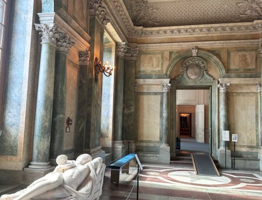Imagen del interior del palacio de estilo neoclásico. En la sala hay una escultura de una mujer amamantando a un niño tumbados. Figuras clásicas de Grecia o Roma.