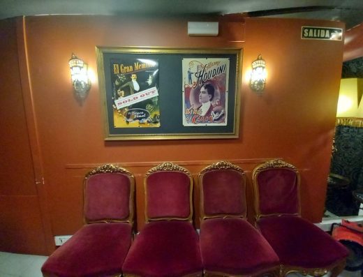 Imagen de la zona de espera en la recepción con varias sillas. Colgado en la pared hay un cuadro con varios carteles de la obra del mago que indican entradas vendidas.