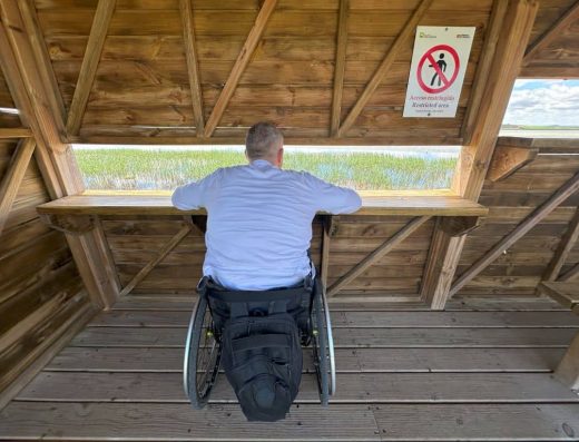 Imagen de una persona en silla de ruedas junto a la ventana del mirador observando el paisaje de la laguna.
