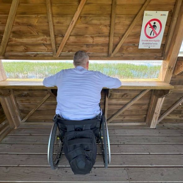 Imagen de una persona en silla de ruedas junto a la ventana del mirador observando el paisaje de la laguna.
