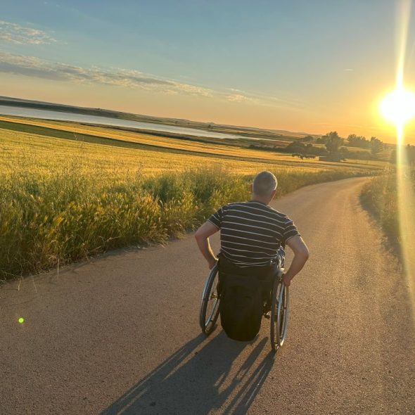 Imagen de una persona en silla de silla de ruedas recorriendo un sendero junto a la laguna al atardecer.