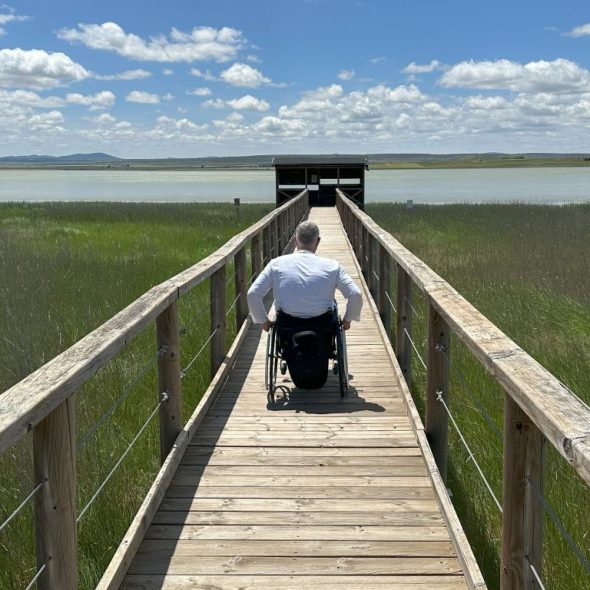 Imagen de una persona en silla de ruedas accediendo por la pasarela de madera hacia el mirador de la laguna.