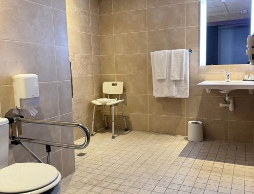 Imagen general del baño con lavabo sin pie, ducha a cota cero, silla de ducha e inodoro con barra de apoyo.
