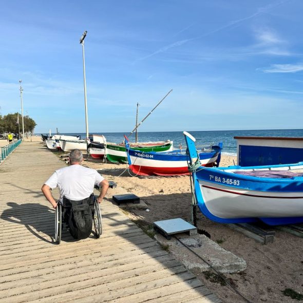 Imagen de una persona usuaria de silla de ruedas recorriendo un sendero de madera junto a unas pequeñas embarcaciones en la playa