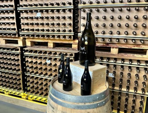 Imagen de una barrica con botellas sobre ellas. Detrás botelleros.