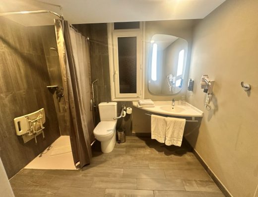 Imagen del interior del baño adaptado. Inodoro con barras, ducha a cota cero con asiento. y lavabo sin pie.