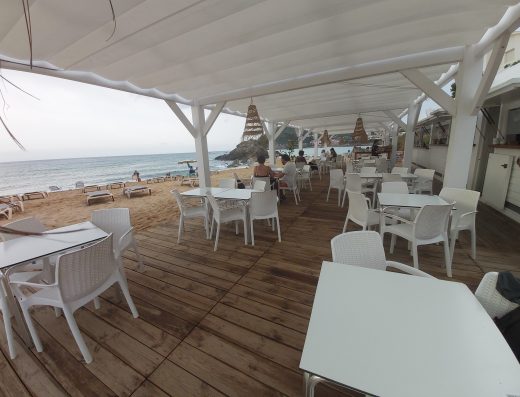 Imagen de la terraza de un chiringuito con varias mesas con gente. Situado junto a la playa donde se observan numerosas hamacas.