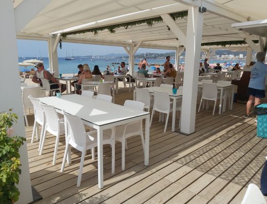 Imagen de la terraza de un chiringuito con abundante gente. Situado junto a la playa. Numerosos barcos navegan junto a la costa.