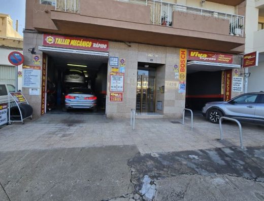 Imagen de la fachada de un edificio con tres entradas. La entrada de la derecha y de la izquierda son entrada a un taller mecánico. En medio de ambas hay la entrada a un portal.