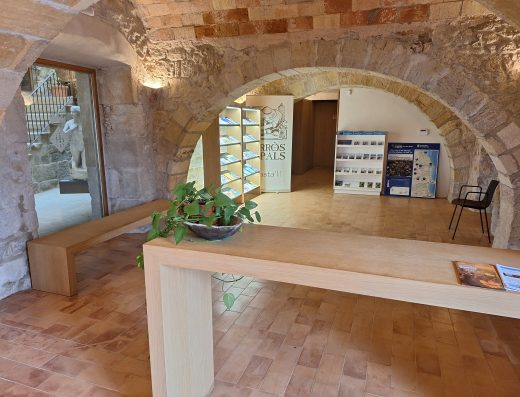 Imagen del interior de la oficina de turismo con arcos de medio punto que dividen los espacios. Las paredes son de piedra. En primer plano un mostrador. A la izquierda un banco de descanso y en un segundo plano varios dispensadores de folletos.