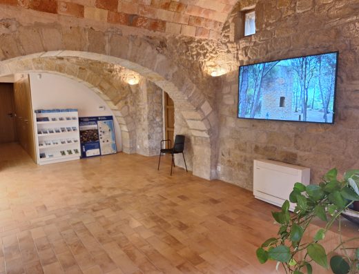 Imagen del interior de la oficina de turismo con muros de piedra y arcos de medio punto. En la pared hay una pantalla donde se ve una torre circular entre altos árboles.