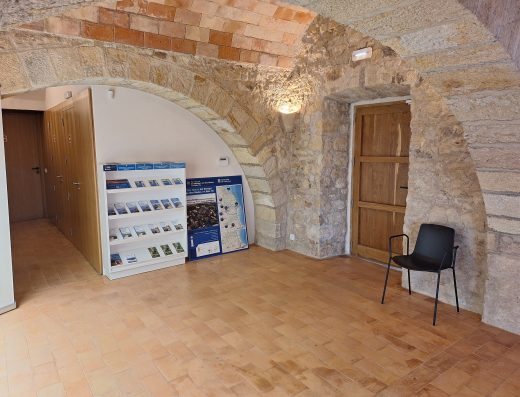 Imagen del interior de la oficina de turismo con muros de piedra y arcos de medio punto. Al fondo lun expositor con folletos turísticos. En la derecha de la imagen hay una puerta de madera y junto a ella una silla.