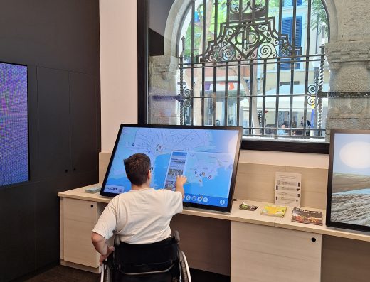 Imagen del interior de una oficina de turismo donde hay una una usuaria de silla de ruedas tocando una pantalla interactiva.