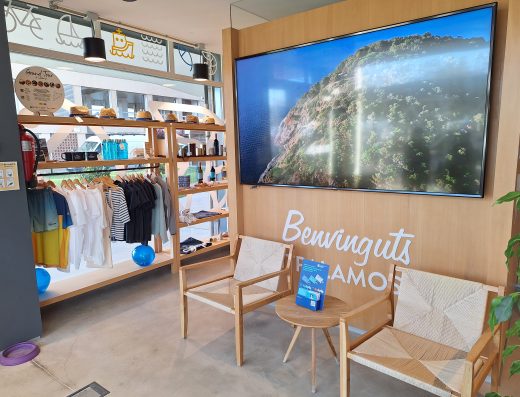 Imagen del interior de la oficina de turismo con un expositor en el escaparate con distintos productos promocionales como camisetas, sombreros, botellas, etc. Junto a él una zona de espera con con dos sillas y una mesita redonda entre ellas. En la pared una imagen aérea de la costa escarpada.
