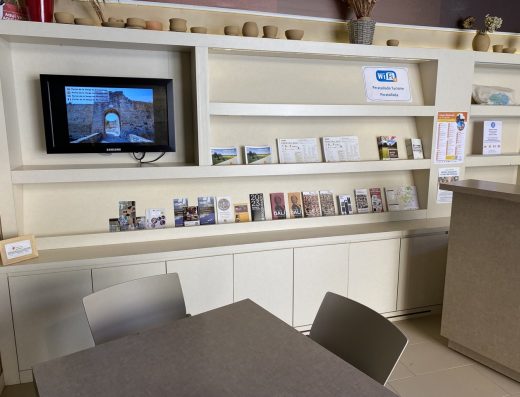 Imagen del interior de una oficina de turismo con mobiliario con estanterías donde se colocan los folletos informativos. En uno de los huecos hay una pantalla. Delante del mostrador hay una mesa con sillas.