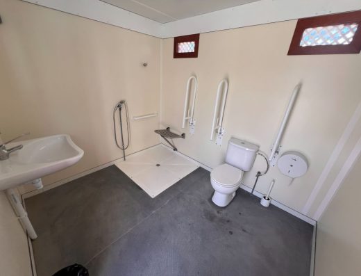 Imagen del interior de un baño adaptado con inodoro con barras de apoyo abatibles en ambos lados. A la izquierda está la ducha a cota cero con asiento y frente a ella el lavabo sin pie.
