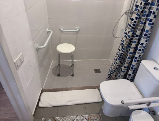 Imagen del interior del baño. A la derecha de la imagen el inodoro con una barra de apoyo y al fondo la ducha con taburete junto a las barras de apoyo.
