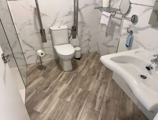Imagen de un inodoro con barras de apoyo abatible a ambos lados. A la derecha de la imagen, en primer plano, hay un lavabo sin pie.