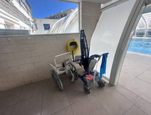 Imagen de una silla de ruedas de agua y una grúa hidráulica portátil junto a la puerta de entrada a la zona de las piscinas.
