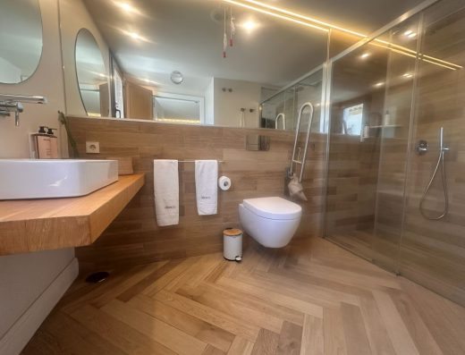 Imagen del interior del baño adaptado. En el centro el inodoro con barra de apoyo, a la derecha la ducha con mampara y a la izquierda el lavabo sin pie.