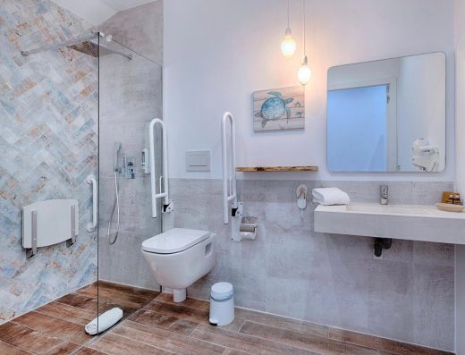 Imagen del interior del baño adaptado. A la izquierda la ducha con mampara y asiento. En el centro de la imagen, junto a la ducha, el inodoro con barras de apoyo abatibles a ambos lados. A la derecha el lavabo sin pie.