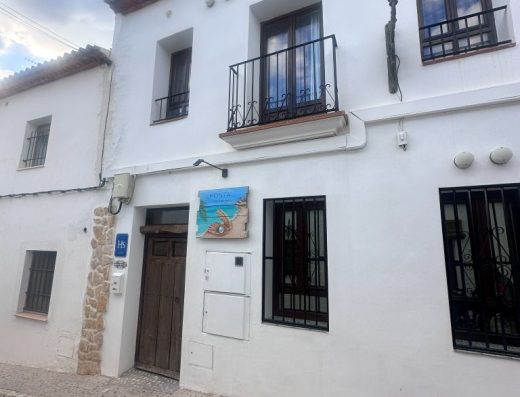 Imagen de la fachada principal del hostal. Edificio de dos alturas con pared blanca. La calle es empedrada. El cartel del hostal tienen un dibujo de una concha con una perla blanca sobre la arena de la playa junto a la orilla.