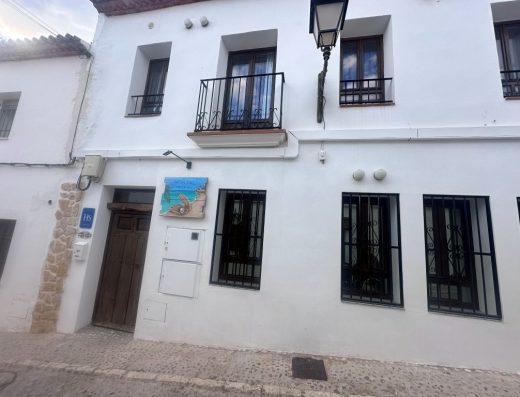 Imagen de la fachada principal del hostal. Edificio de dos alturas con pared blanca. La calle es empedrada. El cartel del hostal tienen un dibujo de una concha con una perla blanca sobre la arena de la playa junto a la orilla.
