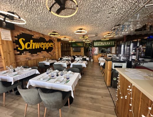 Imagen del interior del restaurante. Las mesas del salón están preparadas con la vajilla y cubertería. EN la pared de la izquierda hay un gran cartel en el que se lee Scheweppes y al fondo otro en el que pone Restaurante.