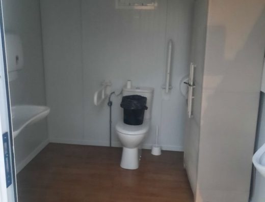 Imagen del interior de un aseo adaptado con inodoro con barras de apoyo abatibles en ambos lados. A la derecha del inodoro hay un armario.