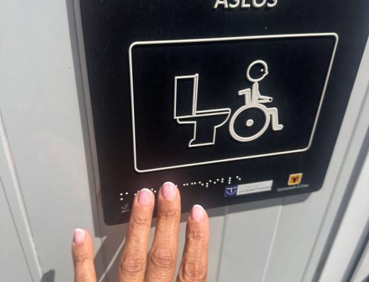 Imagen de una mano tocando un pictograma en relieve y braille de un aseo adaptado con símbolo de accesibilidad.
