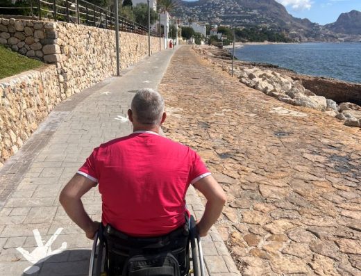 Imagen de una persona en silla de ruedas recorriendo el sendero sobre pavimento de hormigón impreso. A la derecha se encuentra el mar.