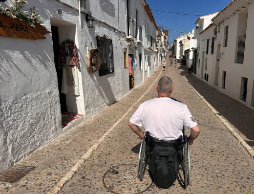 Imagen de una persona usuaria de silla de ruedas transitando por medio de la calle. La calle tiene pavimento empedrado con aceras con pequeño bordillo y casas.