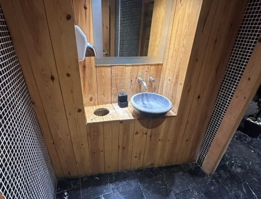 Imagen del lavabo ubicado en el exterior de la cabina de aseo. Está colocado sobre una repisa de madera, sin hueco inferior libre.