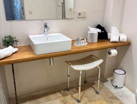 Imagen de un lavabo con una balda de madera que lo sostiene que va de lado a lado de la pared. Debajo de hay un taburete para la ducha.