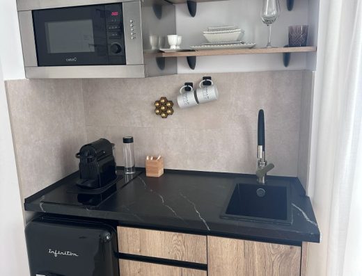 Imagen de un pequeño espacio de cocina con una vitrocerámica y un pequeño lavabo. En la parte superior hay un microondas a la izquierda y varias baldas a la derecha donde se han colocado una jarra, un tostador y algún vaso.