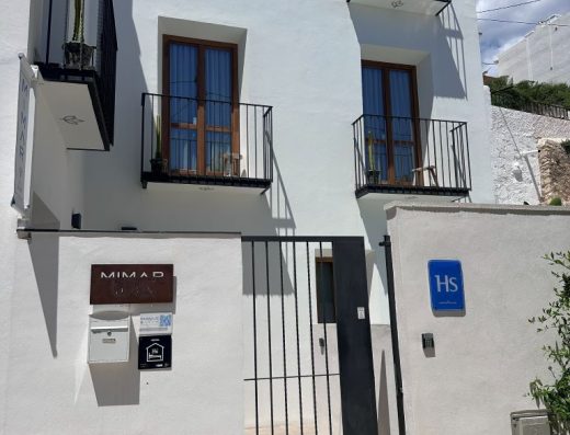 Imagen de la entrada al hotel con una puerta enrejada al recinto y posteriormente el edificio con fachada blanca.