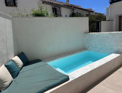 Imagen de una pequeña piscina exterior elevada. En el lado izquierdo hay una colchoneta con cogines.