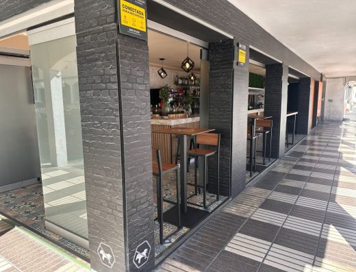 Imagen del exterior del local con fachada abierta donde están colocadas unas mesas altas con taburetes.