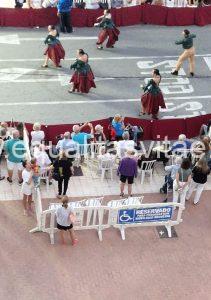 Imagen de un grupo de personas desfilando con trajes tradicionales. En la acera hay un espacio reservado para personas con movilidad reducida. En el hay numerosas sillas ocupadas, usuarios de andadores y una persona en silla de ruedas.