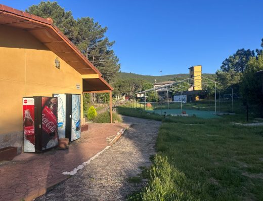 Imagen del exterior con un sendero enlosado junto a una zona ajardinada. A la izquierda el edificio con dos máquinas expendedoras de bebidas junto a la pared.