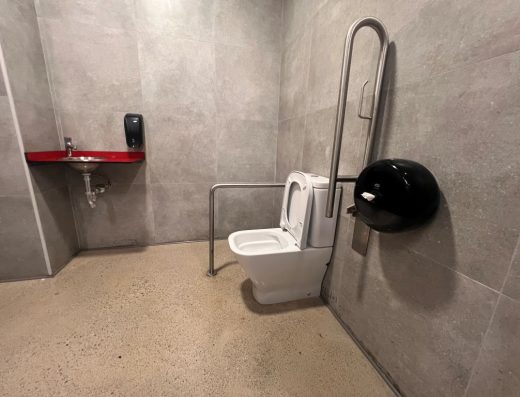 Imagen del interior de un aseo adaptado. A la derecha un aseo con barras de apoyo, al fondo a la izquierda, colocado en la esquina hay un lavabo sin pie.