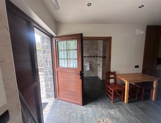 Imagen de la entrada a la casa desde el interior. Puerta abatida hacia adentro. Al fondo, de frente, la entrada al baño. A la derecha una mesa de madera con dos sillas y dos taburetes.